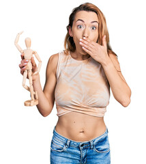 Poster - Young caucasian woman holding small wooden manikin covering mouth with hand, shocked and afraid for mistake. surprised expression