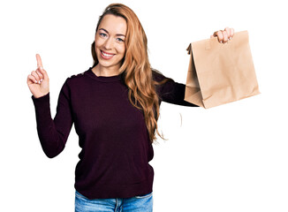 Canvas Print - Young caucasian woman holding take away paper bag smiling with an idea or question pointing finger with happy face, number one