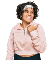 Young hispanic woman with curly hair wearing sportswear smiling with happy face looking and pointing to the side with thumb up.