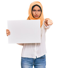 Poster - Young brunette arab woman wearing traditional islamic hijab holding banner pointing with finger to the camera and to you, confident gesture looking serious