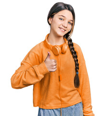 Poster - Young brunette girl with long hair wearing sweatshirt and headphones doing happy thumbs up gesture with hand. approving expression looking at the camera showing success.