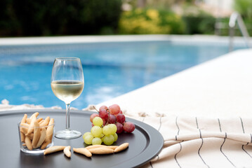 A glass of white wine with snacks on a tray by the pool.