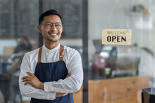 Asian Happy Business Man In Glasses Is A Waitress In An Apron, The Owner Of The Cafe Stands At The Door With A Sign Open Waiting For Customers , Cafes And Restaurants Small Business Concept.