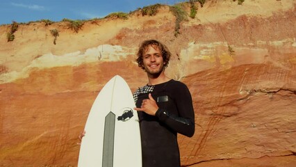 Wall Mural - Young surfer standing at the shore of the beach and showing shaka gesture