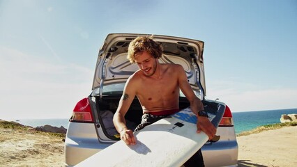 Wall Mural - Sporty shirtless man waxing surfboard at the car trunk on sand beach