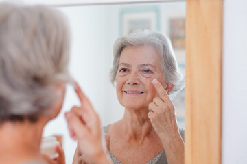 Portrait of senior beautiful woman applies anti aging cream on wrinkled face - elderly smiling lady holding a cosmetic jar product