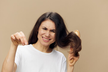 A sad, upset woman combs her hair with a wooden massage comb and screams, showing the fallen hair to the camera. Horizontal photo on a beige background.