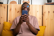 © Daniel - Young black man laughing at home relaxing using mobile phone to watch funny videos. Reading text message.