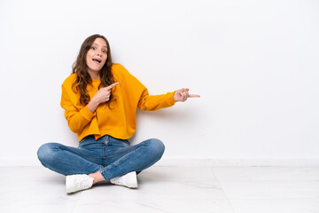 Wall Mural - Young caucasian woman sitting on the floor isolated on white wall surprised and pointing side