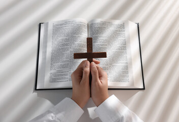 Wall Mural - Above view of woman praying with wooden cross over Bible at white table