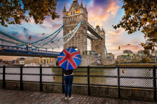 London In Autumn Time Concept With A Tourist Person Holding A British Flag Umbrella In Front Of The Famous Tower Bridge During Sunset Time