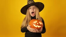 Little Girl In Witch Hat Isolated On Yellow Background. Pretty Blonde Witch Carrying Orange Pumpkin Lantern. Halloween Holiday. Female Laughing With Scary Pumpkin In Hands. Pumpkin With Face. 4K, UHD