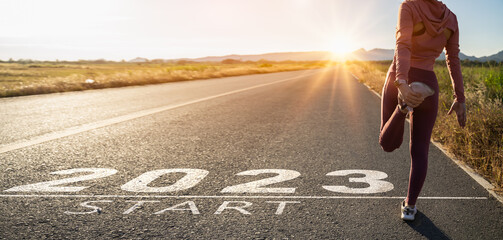 New year 2023 or start straight concept.word 2023 written on the asphalt road and athlete woman runner stretching leg preparing for new year at sunset.Concept of challenge or career path and change.