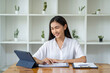 © crizzystudio - An Asian businesswoman wipes a to-do list on a tablet and responds to customer chats at a desk in a modern office.