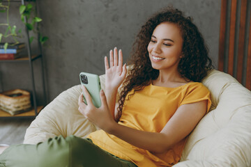 Sticker - Young happy woman of African American ethnicity in casual clothes use mobile cell phone waving hand sit in armchair stay at home flat rest relax spend free spare time in living room indoor grey wall