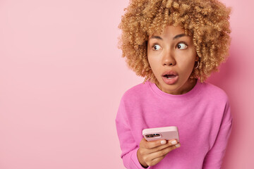 Wall Mural - Indoor shot of scared curly haired adult woman looks shocked aside holds mobile phone finds out shocking news dressed in pullover keeps jaw dropped isolated over pink background empty space.