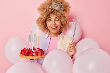 Canvas Print - Surprised cheerful European woman with curly bushy hair holds magic wand strawberry cake withh burning candles celebrates birthday surrounded by inflated balloons isolated over pink background