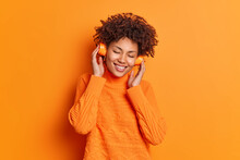 Horizontal Shot Of Satisfied Curly Haired Woman Keeps Hands On Headphones Keeps Eyes Closed From Satisfaction Dressed In Casual Jumper Catches Every Bit Of Music Isolated Over Orange Background