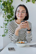 © WHstudio Leushin N - Vertical shot of thoughtful young Asian woman wears transparent eyeglasses and striped jumper drinks coffee looks away sits at white desk with modern technologies around against cozy interior.