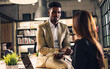 © Ann Rodchua - Handsome African businessman wearing formal suit, seriously talking, discussing business and project plan with colleague, sitting on desk with laptop in modern indoor meeting room at office.