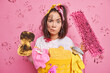 Displeased serious woman frowns face and raises eyebrows focused at camera does washing at home holds dirty sponge and mop poses near basket of collected laundry isolated over pink background
