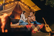 © bondvit - Two Smiling female friends drinking wine and eating fruits sitting in cozy glamping tent in autumn evening bonfire. Luxury camping tent for outdoor holiday and vacation. Lifestyle concept