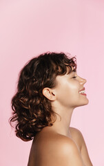 Wall Mural - Profile photo of young female model, curly hair, closed eyes, laughing, advertising of skin care treatment, cosmetic and spa product on pink background