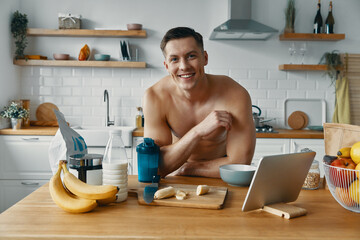 Wall Mural - Confident young man looking at camera and smiling while preparing healthy food at the kitchen