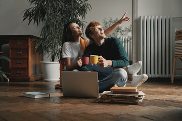 Wall Mural - Dreamful young couple enjoying hot drinks and pointing away while sitting on the floor at home