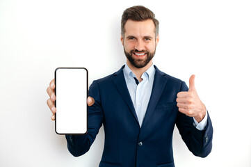 Happy confident caucasian bearded man, in a suit, showing smart phone with empty mock-up screen in hand for advertising, points finger thumb up gesture, standing on isolated white background, smiles