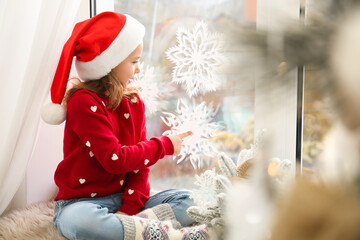 Wall Mural - Little girl in Santa hat near window decorated with paper snowflakes at home