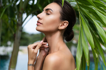 Beauty portrait, soft skin. Female model with natural make-up and healthy skin behind a green leafy plant. Portrait of a beautiful girl with nude nails, bare shoulders