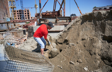 Floor screeding contractor shoveling sand-cement mix outdoors at construction site. Male worker with shovel in his hands preparing cementitious material for floor screed.