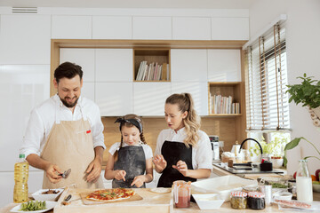 Delighted surprised kid and mother waiting for eating homemade domestic pizza. Father jolding pizza cutter cutting pizza. Happy family preparing cooking baking dinner in modern kitchen with window.