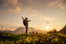 Mature Man Standing With Arms Outstretched On Top Of Mountain At Sunset