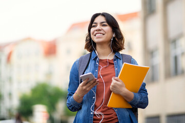 cheerful young arab student female listening music with smartphone and earphones outdoors
