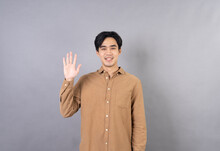 A Happy, Smiling Young Asian Man Gives A Waiving Saying Hello With Five Fingers Up In Greeting Body Language, Studio Photo, Friendly Welcome Gesture, And Happiness.