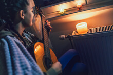 Teenage Girl Sits Under Blanket Near Heating Radiator With Candles And Play Guitar .Rising Costs In Private Households For Gas Bill Due To Inflation And War, Energy Crisis