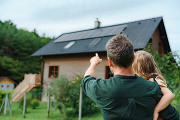 rear view of dad holding her little girl in arms and showing at their house with installed solar pan