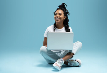Happy young woman sitting on the floor with crossed legs and using laptop on blue background.