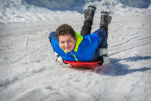 In Winter, In A Forest, A Boy Lying On His Stomach Slides Down A Hill On A Plastic Plate. He Is Joyful.
