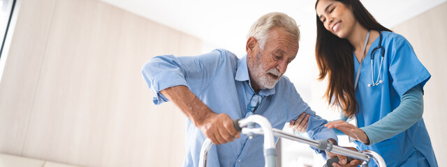 Portrait of Asian young nurse helping old elderly disable man grandfather to walk by using walker equipment in the bedroom. Senior patient of nursing home moving with walking frame and nurse support