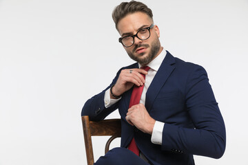 Wall Mural - arrogant young businessman sitting on chair and adjusting red tie