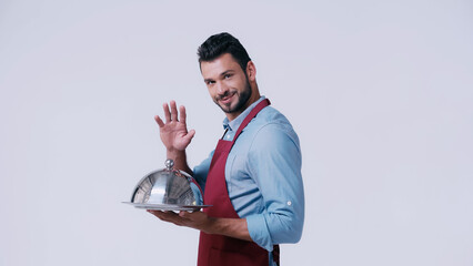 Poster - positive waiter waving hand while holding serving dish with cloche isolated on grey.