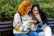 Two woman friends sitting on bench in public park with mobile phone on hand, talk and smile