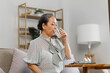 © NanSan - Senoir mature asian woman with glass of water taking pills, sitting on couch.