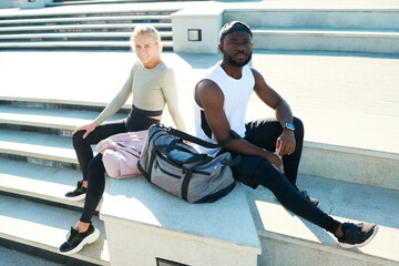 Wall Mural - Young intercultural man and woman in sportswear sitting by concrete staircase in front of camera in urban environment in the morning