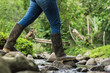 medium shot of a peasant girl crossing a stream in black marsh boots, stepping on the stones to avoid falling into the water. green background of rainforest in summer.