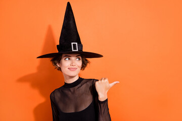 Wall Mural - Photo portrait of charming young girl offer look empty space promo dressed trendy black halloween witch look isolated on orange background