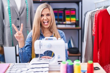 Sticker - Blonde woman dressmaker designer using sew machine smiling with happy face winking at the camera doing victory sign with fingers. number two.